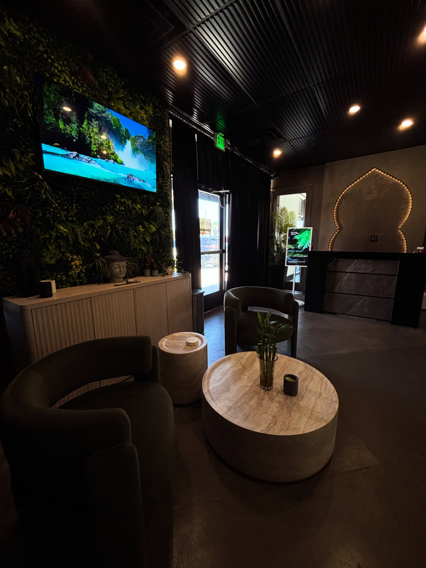 Front lobby and waiting lounge at The Temple Head Spa in Las Vegas with a living plant wall, Buddha statue, sculptural chairs, travertine tables, and an illuminated carved arch above the reception desk
